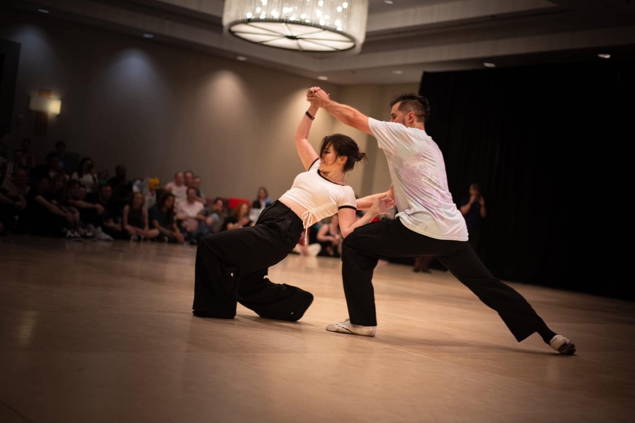 Kristen mid-dance with a partner on a ballroom floor beneath a chandelier, leaning away in a stretched two-hand connection