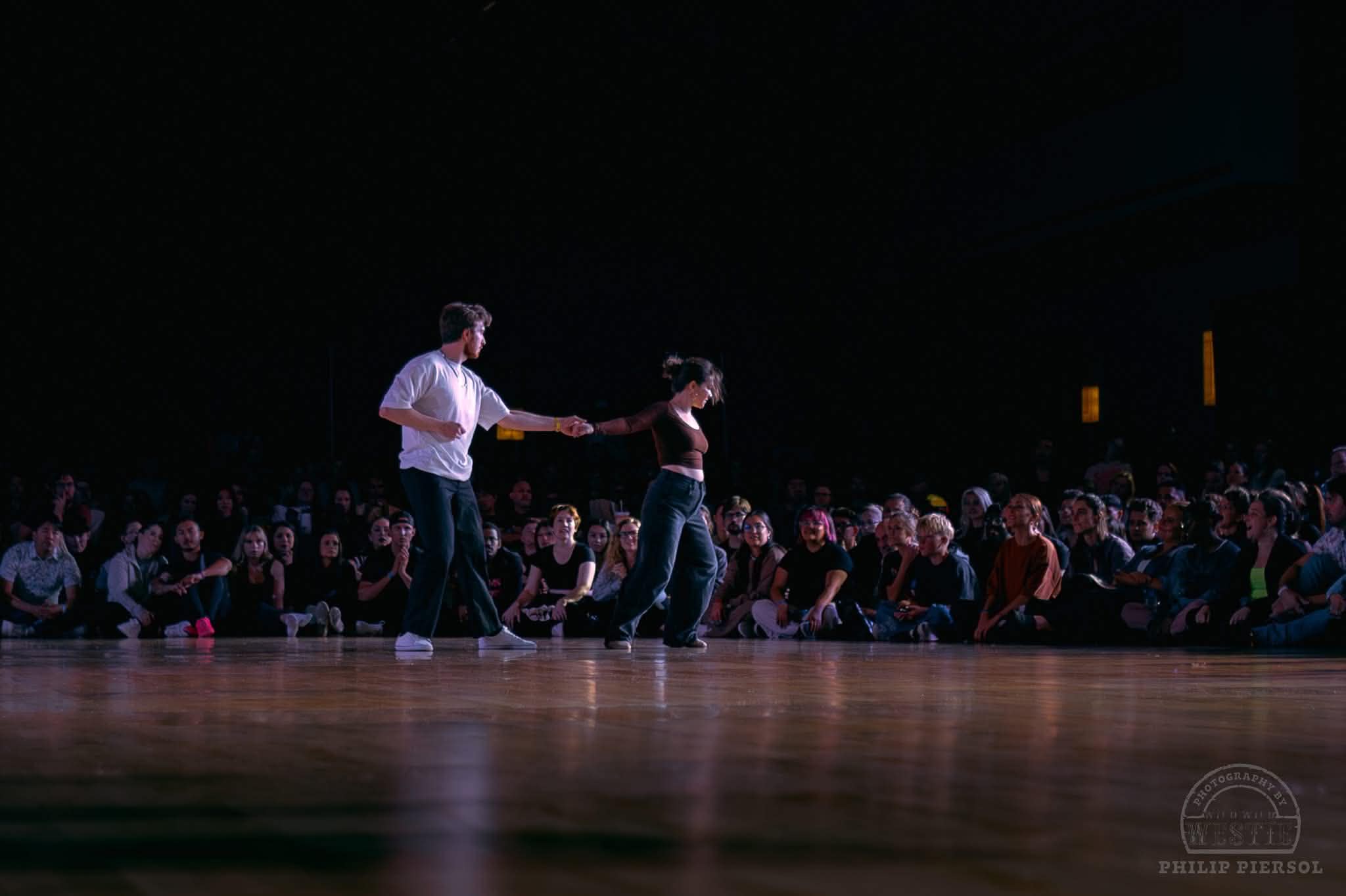 Two dancers in a spotlight surrounded by a seated crowd at a West Coast Swing event